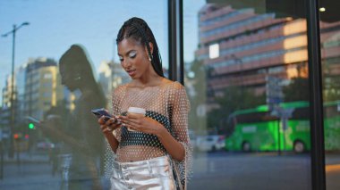 Koyu renk deri modeli, cam bina duvarında akıllı telefon yazıyor. Modayı takip eden modacı cep telefonuyla selfie çekmek için elinde kahve tutuyor. Yaratıcı makyajlı esmer kadın cep telefonu kamerası pozu veriyor.