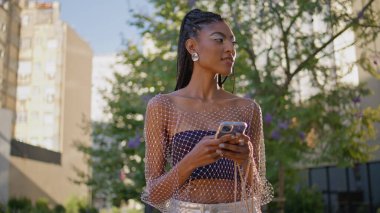 Yaz sokağı kapanışında akıllı telefonlara bakan neşeli kız. Neşeli Afro-Amerikalı kadın şehir merkezindeki modern cep telefonuyla mesaj atıyor. Moda kıyafetleri içinde cep telefonu cihazını tutan mutlu kadın.
