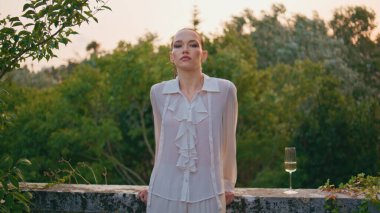 Moda kızı, şarap kadehi görünümlü kamerayla dışarıda poz veriyor. Beyaz tişörtlü sakin bir kadın balkonda duruyor ve sakinliğini hissediyor. Gerçekten çekici bir kadın yeşil ağaçların önünde çitlere yaslanıyor.