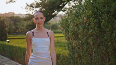 Yazın manzarada yürüyen zarif bir kız. Gülümseyen genç kadın kameraya bakıyor, patikada geziniyor, tek başına güzel bir bahçe. Beyaz elbiseli muhteşem mutlu bayan yeşil parkta çalılarla dinleniyor.
