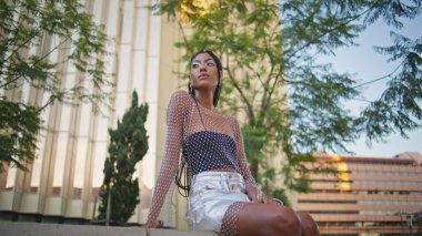 Afro örgülü kadın şehir meydanındaki bankta erkek arkadaşını bekliyor. Yalnız Afrikalı Amerikalı kadın yeşil ağaçlar parkında dinleniyor ve yaz havasının tadını çıkarıyor. Moda kıyafeti içinde kahverengi deri modeli dinleniyor.