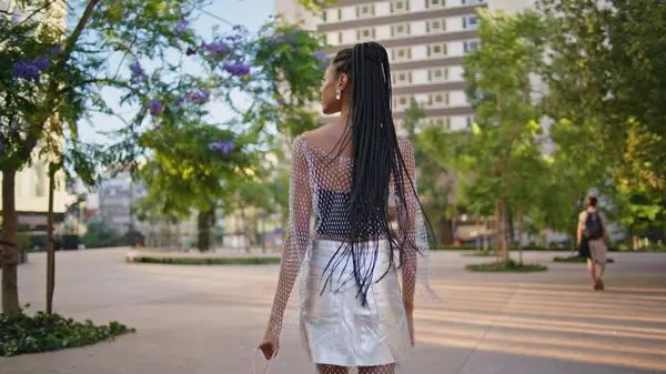 Relaxed brunette walking city back view. Tourist african american girl enjoying summer stroll looking asides at downtown. Stylish fashionista with afro braids hairstyle stepping square on high hills