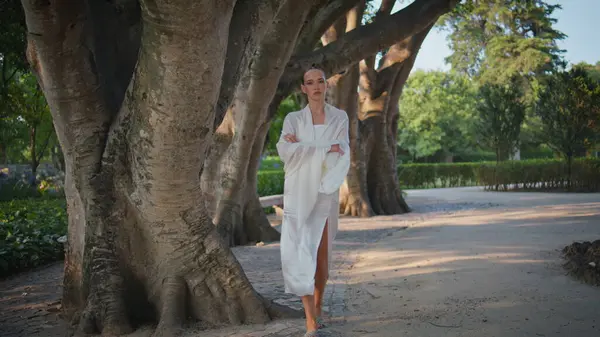 Relaxed woman strolling beautiful city park summer day. Romantic attractive girl walking under wide trees alley enjoying calm weekend. Stylish serious lady wrapped in white cloth relax alone at nature