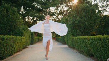 Rüzgarda sallanan beyaz eşarplı güzel yeşil bahçe yolunda yürüyen sakin bir kadın. Gülümseyen zarif bayan antika mermer heykellerle patika parkına gidiyor. Rahatlamış kaygısız kız doğada geziniyor..