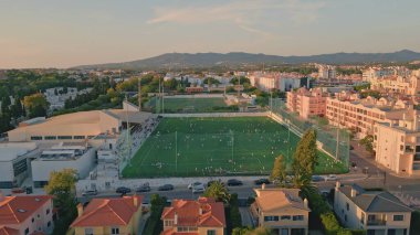 Drone Manzaralı küçük kasaba stadyumu yumuşak akşam güneşi altında. Hafta sonları dinlenen insanlarla dolu güzel bir futbol sahası. Arka planda dağlar olan sakin bir şehir. Sıcak kasaba mahallesi.