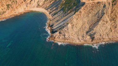 Turkuaz okyanus suyu drone manzaralı güzel, güneşli sahil uçurumları. Sersemletici deniz dalgaları sert taş sahilleri çok yavaş çekimde yıkıyor. Yaz akşamında boş kayalık kıyı şeridi. Harika bir sahil