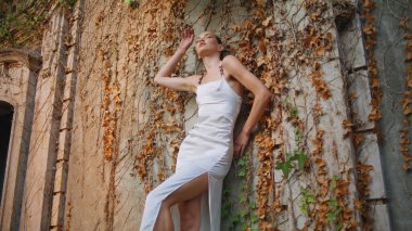 Şehvetli bir kadın duvarda poz veriyor. Sarı sonbahar yapraklarıyla kaplanmış. Zarif beyaz elbiseli seksi güzel manken eski bina kaldırma koluna yaslanıyor. Terk edilmiş antika sarayda duran şık moda kızı..