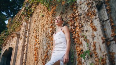 Ortaçağ binasına yaslanan rahat kadın sonbahar yapraklarını kapladı. Eski duvarda beyaz elbiseli, terk edilmiş sarayda poz veren kendine güvenen güzel bir manken. Çekici bir kız kameraya bakıp şehvetini gösteriyor..