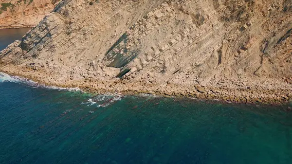 Hava manzaralı güneşli kayalık kıyı şeridi turkuaz okyanus dalgalarıyla süper yavaş çekimde yıkanır. Sakin mavi deniz suyunun kumlu sahil kayalıklarına yuvarlandığı inanılmaz bir deniz manzarası. Resimli yaz deniz manzarası.