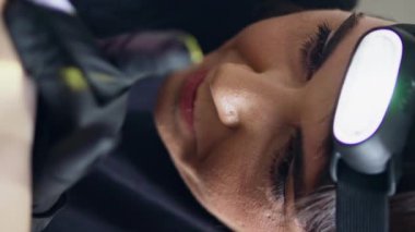 Salon dikey portresinde dövme makinesi kullanan ciddi bir sanatçı. Lateks siyah eldiven dövmecisi ev stüdyosunda çalışıyor. Odaklanmış kadın, el feneri takan müşterinin vücuduna resim çiziyor. Sanat konsepti