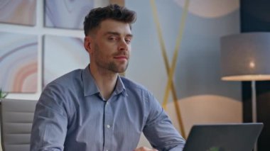 Sakin adam dizüstü bilgisayarlı ofis masasına yakın oturmaktan hoşlanıyor. Gülümseme portresi olan rahat iş adamı görünümlü bir kamera. Genç kendine güvenen başlatıcı görünüm uzaklığı bilgisayar işyerinde dinlendirici