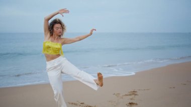 Sahilde dans eden koreograf esnek vücutlu. Kıvırcık profesyonel dansçı kadın akşam okyanus kıyısında modern stilde pratik yapıyor. Genç şehvetli kız deniz kıyısında doğaçlama dans ediyor..