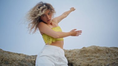 Gökyüzüne karşı dans eden etkileyici kadın doğaya modern koreografi sergiliyor. Yakından. Çekici kıvırcık kız taş kayalarda şehvetle hareket ediyor. Kameraya bakıp düzgün hareketler yapan sarışın.