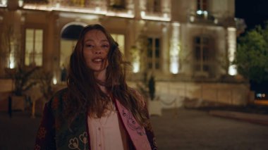 Akşam kentinde poz veren güzel bayan kameraya yakından bakıyor. Gece kasabasında lüks bir binanın önünde duran güzel, gülümseyen bir kadın. Neşeli mutlu turist kız saçlarını sallayan rüzgara dokunuyor. Gece hayatı konsepti.
