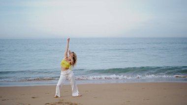 Kız okyanus kıyısında modern dans gösterisi yapıyor. Kum plajında çağdaş abartılı koreografi uygulayan çekici kadın dansçı. Kıvırcık şehvetli koreograf cesedi boş deniz kıyısında taşıyor..