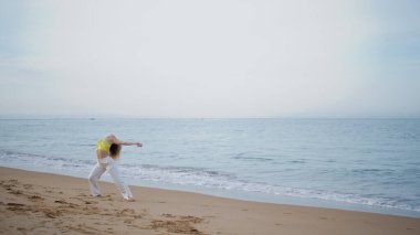 Sahilde ıslak kuma basarak dans eden baştan çıkarıcı kadın. Okyanus dalgaları yakınında çağdaş koreografi pratiği yapan güzel bir kız dansçı. Kıvırcık kadın sanatçı plajda modern serbest stil performans sergiliyor..
