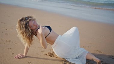 Kum güneşli sahilde dans eden bir kız. Yakın çekim. Şehvetli genç bir kadın, ıslak deniz kıyısında, yaz akşamında sorunsuzca hareket ediyor. Kıvırcık profesyonel dansçı okyanus kıyısında doğaçlamanın keyfini çıkarıyor..