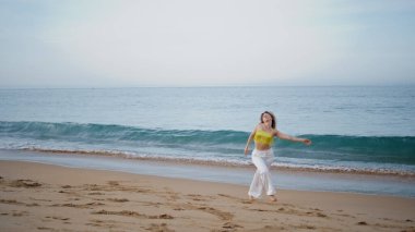 Kum plajında modern tarzda doğaçlama dans eden şehvetli kız. Sahilde çağdaş koreografiyi icra eden zarif genç bir kadın. Kıvırcık kadın sanatçı vücudu okyanus dalgalarının önünde hareket ettiriyor..