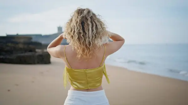 Serene woman strolling beach touching curly hair back view. Unrecognizable girl traveler stepping on sand seashore enjoy summer vacation. Carefree lady walking beautiful ocean coast looking seascape