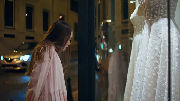 Girl looking at wedding store showcase standing on night city street close up. Attractive woman interested by expensive shiny bridal dress on shop mannequin. Future bride admiring white luxury gown.