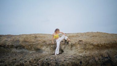 Serbest stil dansçısı taşlı kayalarda dans ediyor. Güzel kadın sanatçı esnek vücudu doğada moda kıyafetlerle eğiyor. Kıvırcık kadın sanatçı açık havada mavi gökyüzüne karşı modern koreografiyle dans ediyor.