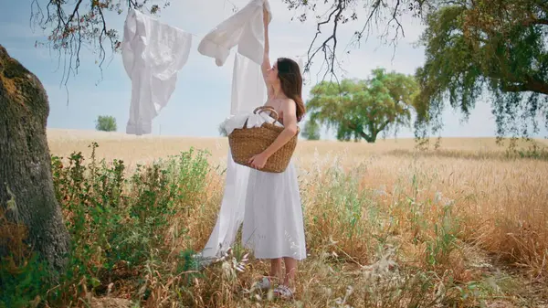Country lady taking dry linen from rope at summer field. Young brunette put clean washed laundry to wicker basket at countryside nature alone. Tender calm woman in courtyard. Village lifestyle concept