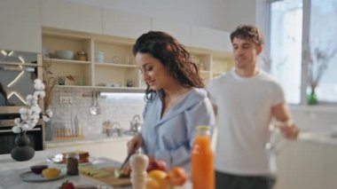 Koca, sıcak bir apartman mutfağında genç eşine kahvaltı hazırlamaya yardım ediyor. Sağlıklı öğle yemeği için meyve dilimleyen mutlu, gülümseyen kadın. Neşeli adam meyve suyu sarılması için gözlük alıyor..