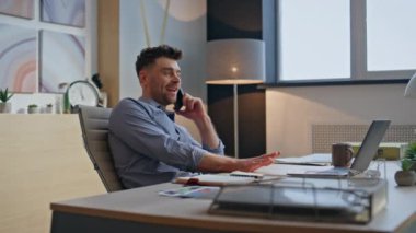 Cep telefonuyla konuşan mutlu bir adam akşamları yakın planda oturuyor. Gülümseyen pozitif iş adamı telefon görüşmesine gülüyor yaratıcı ajansta dizüstü bilgisayar masasında çalışıyor. Neşeli işçi tek başına akıllı telefonla konuşuyor. 