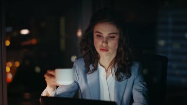 Karanlık işyerinde kahve içen gece çalışanı laptopa yakından bakıyor. Geceleri kurumsal bilgisayarda çalışan ciddi yorgun kadın yönetici. Fazla mesai yapan iş kadını..