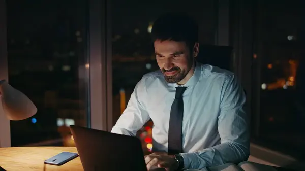 Enthusiastic worker sitting dark office working on laptop with smile close up. Motivated bearded businessman passionate about project implementation at workplace late evening. Happy workaholic smiling