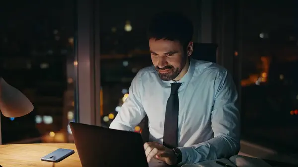 Male workaholic working late evening on laptop close up. Enthusiastic businessman looking on computer monitor sitting dark office alone. Confident bearded corporate manager busy by overtime project.