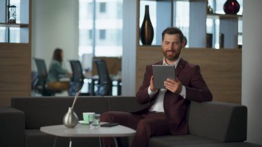 Gülümseyen müdürün rahat ofis binasında gülen bir videosu var. Mutlu sakallı iş adamı, açık alanda tablet bilgisayar ekranını izliyor. Modern emlakçı iş mutluluğu kavramı