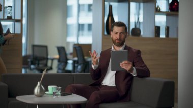 Neşeli patron ofis içi kabinesinde iş videosu konferansından bahsediyor. Mutlu sakallı girişimci rahat koltukta tanımadığı insanlarla tablet bilgisayardan konuşuyor. Dijital adam çalışma konsepti.