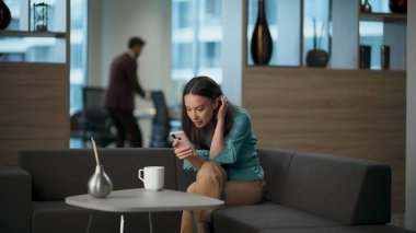 Ofis kanepesinde akıllı telefon izleyen mutlu kadın. Heyecanlı yönetici cep telefonu ekranına bakıyor ve tek başına seviniyor. Gülümseyen CEO, lobide iyi haberler okuyarak başarıyı kutluyor. İş kadını kavram geliştiriyor