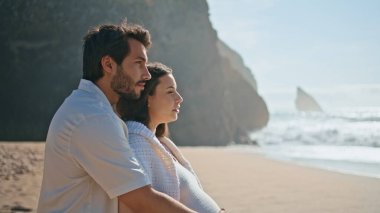 Hamile çift deniz manzarasına bakıyor yaz tatilinde kumsalda dikiliyorlar. Mutlu, rahat eşler bebek için güneşli deniz kıyısında hafta sonu tatili bekliyor. Yakışıklı, sevgi dolu bir adam karısına sarılıyor..