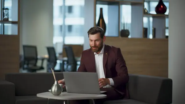 Surprised man staring computer screen at modern coworking interior. Smiling bearded businessman rejoicing work achievement drinking coffee cup. Happy excited manager feeling success winning victory