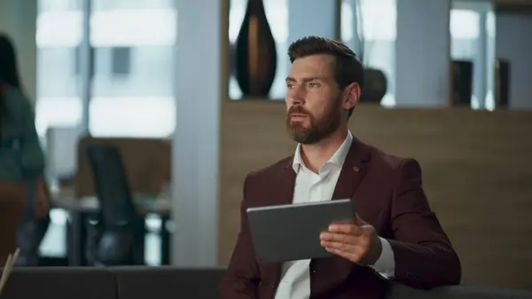 Sad millioner holding tablet computer in comfortable office interior cabinet. Tired bearded professional realtor looking pad screen corporate hall. Man entrepreneur stressful marketing work concept.