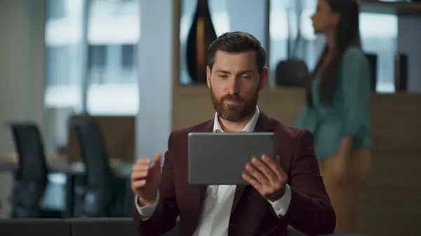Positive business entrepreneur drinking coffee having videocall meeting at office interior hall. Professional successful lawyer making tablet device conference. Bearded realtor holding pad at cabinet