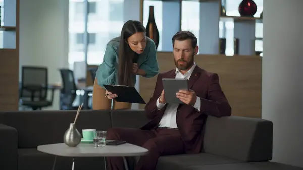 Serious business partners discussing tablet corporate work in big modern office interior. Focused professional lawyer man looking computer screen at comfortable sofa. Successful marketing pair concept