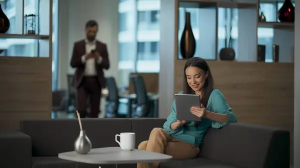 Happy businesswoman watching tab computer at office lobby. Cheerful entrepreneur using tablet with funny emotions laughing. Excited executive scrolling tech device application resting couch alone