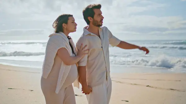 Relaxed future parents walking picturesque ocean coast enjoying sunny landscape. Happy pregnant woman hugging beloved husband strolling beach. Smiling couple expecting baby looking summer seascape.