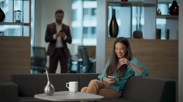 Happy businesswoman watching mobile phone at office lobby. Cheerful entrepreneur using smartphone with funny emotions laughing. Excited executive scrolling tech device application resting couch alone