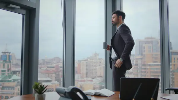 Serious manager enjoy coffee break standing at window modern office. Focused bearded business man drinking espresso at luxury workplace. Entrepreneur standing near work desk holding cup hot beverage.
