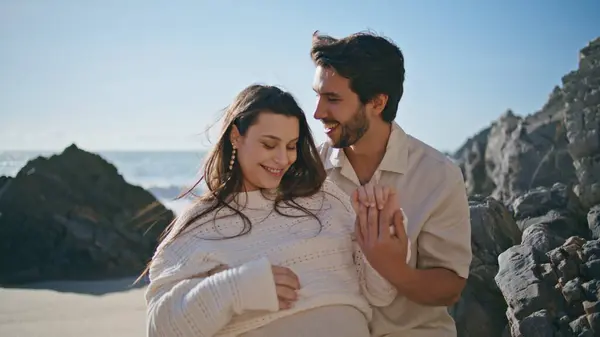 Young couple enjoying pregnancy relaxing on sunny ocean coast at summer weekend close up. Beautiful pregnant woman feeling calmness in gentle husband hug. Happy future parents standing on sandy beach.