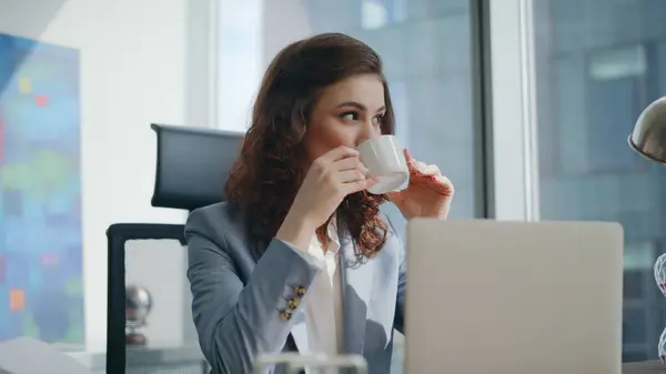 Businesswoman enjoy coffee break sitting modern office desk close up. Calm relaxed woman manager drinking beverage at company workplace looking laptop. Attractive lady employee working at computer.