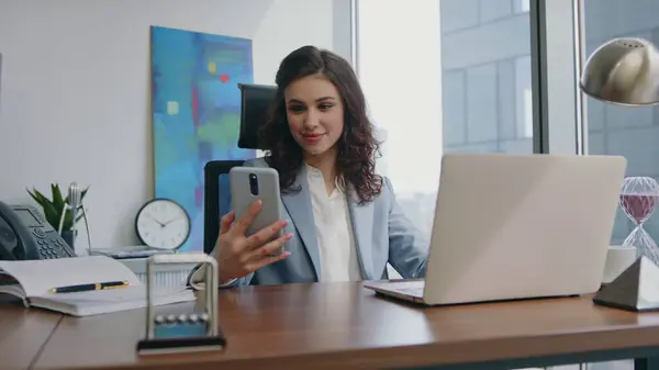 Confident girl looking on smartphone screen feeling surprised at office desk close up. Attractive businesswoman rejoicing phone call smiling in modern interior. Cheerful lady talking telephone happily