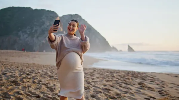Expectant mother video call by cellphone at evening beach. Happy pregnant woman greeting on smartphone camera walking at sand seashore. Smiling girl expecting baby enjoy distant communication on coast