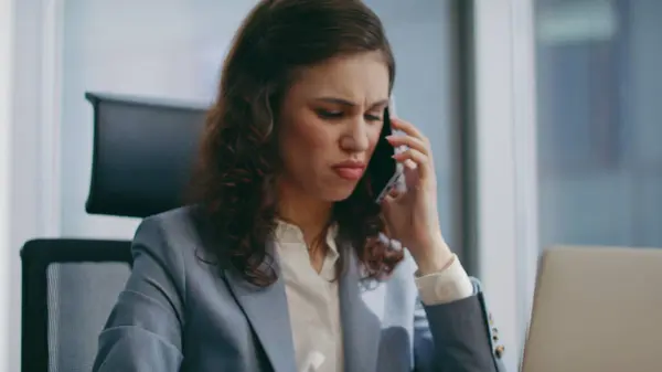 Furious businesswoman shouting in telephone conversation sitting company office close up. Mad young girl manager throwing smartphone getting angry by phone call. Emotional brunette feeling stressed.