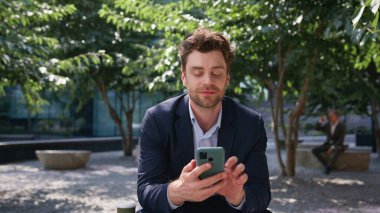 Gülümseyen işadamı cep telefonuna mesaj atıyor. Yeşil güneşli karede tek başına oturuyor. Yaklaş. Zarif takım elbiseli rahat bir yönetici açık havada cep telefonu mesajları okuyor. Akıllı telefon ekranında yazan mutlu yakışıklı adam..