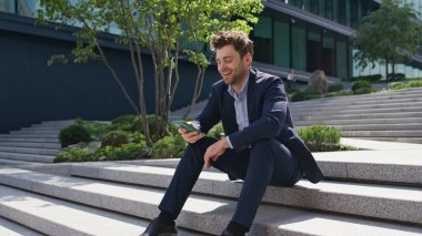 Rahatlatıcı iş adamı akıllı telefonuyla sosyal medyada geziniyor sokak merdivenlerinde zum yapıyor. Mutlu adam cep telefonu ekranına bakıp dışarıda gülüyor. Gülümseyen müdür mesajlaşması ofis dışında iş molası veriyor..